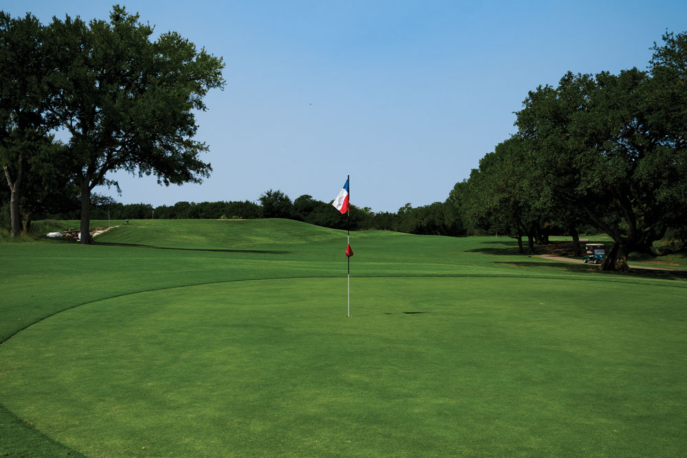 A manicured golf green at Delaware Springs Golf Club with a flagstick topped by a red, white, and blue flag stands in the center—an ideal stop on your Burnet County guys weekend or 3 day itinerary. Trees border the course under a clear blue sky.