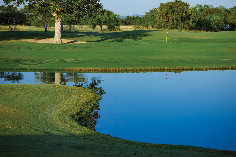 A scene from Legends Golf Course in Kingsland: a green, sand bunker, flagstick, and calm pond reflecting nearby trees and sky. Grass is neatly mowed, and trees are scattered in the background under a clear sky.
