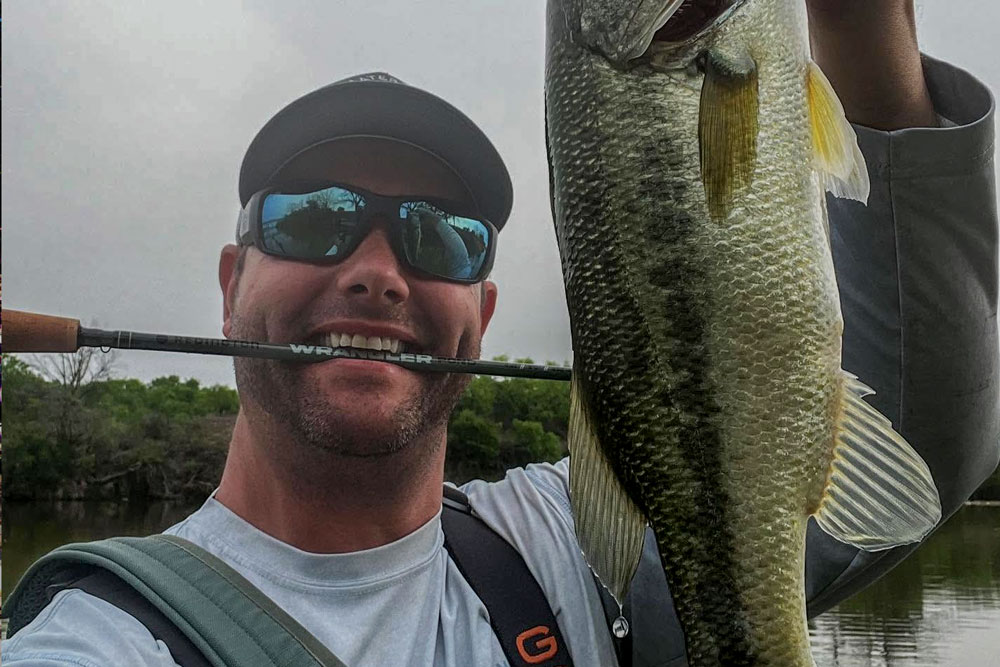 A man wearing sunglasses and a cap smiles while holding up a largemouth bass with one hand and gripping a fishing rod in his mouth, standing near a lake—enjoying another great day with WaterTied Fly Fishing Outfitters.