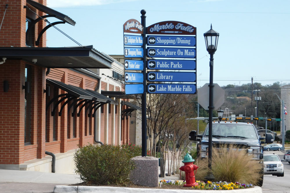 A blue and white directional sign in Marble Falls, Texas, welcomes visitors planning a Girls Weekend or 3 Day Itinerary in Burnet County, pointing to shopping, dining, parks, the library, and Lake Marble Falls amid city sights.