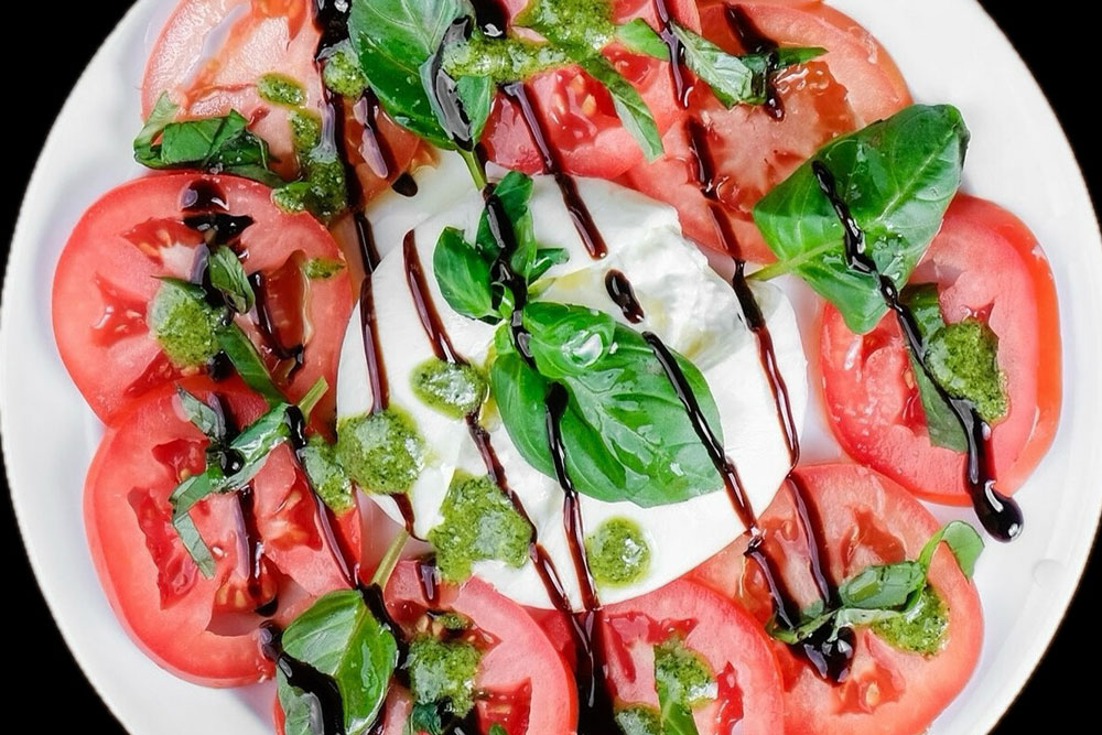 A plate of Caprese salad with sliced tomatoes, fresh mozzarella, basil leaves, and drizzles of balsamic glaze and pesto, beautifully arranged on a white plate at Dalla Tor Restaurant.