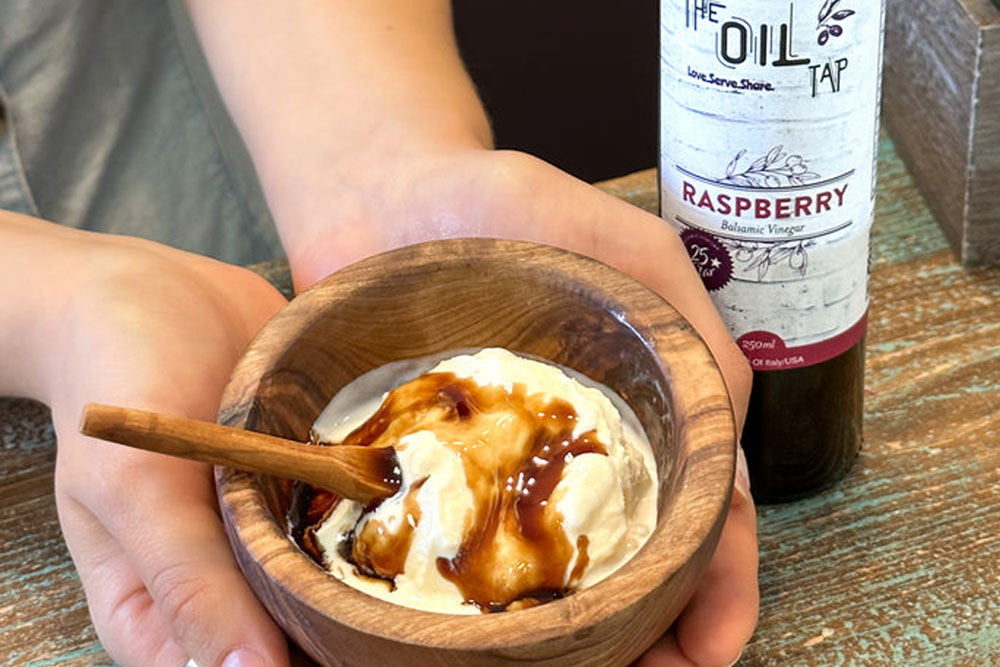 A person holds a wooden bowl of vanilla ice cream drizzled with Oil Tap Marble Falls Raspberry Balsamic Vinegar, with a small wooden spoon inside. A matching bottle stands on a wooden table nearby.