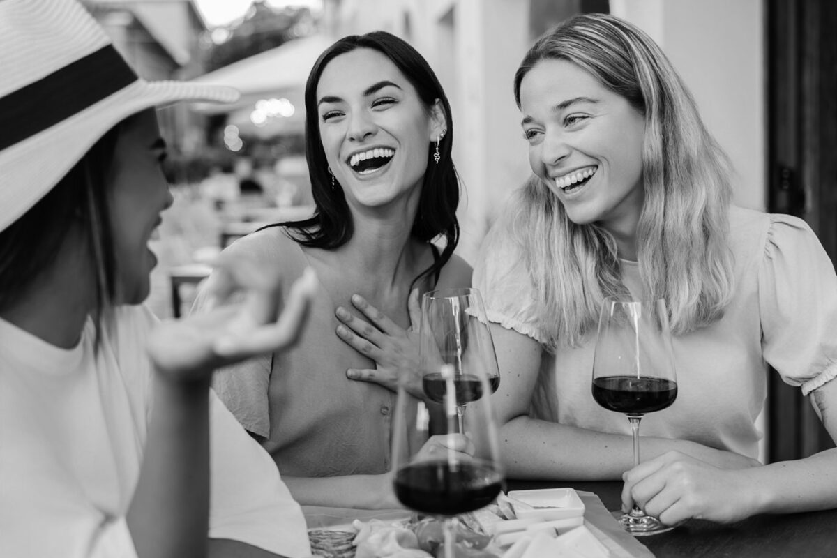 Three women sit at an outdoor table, smiling and laughing together during a Girls Trip in Burnet County. Two glasses of red wine and a plate of snacks are in front of them. The image is in black and white.