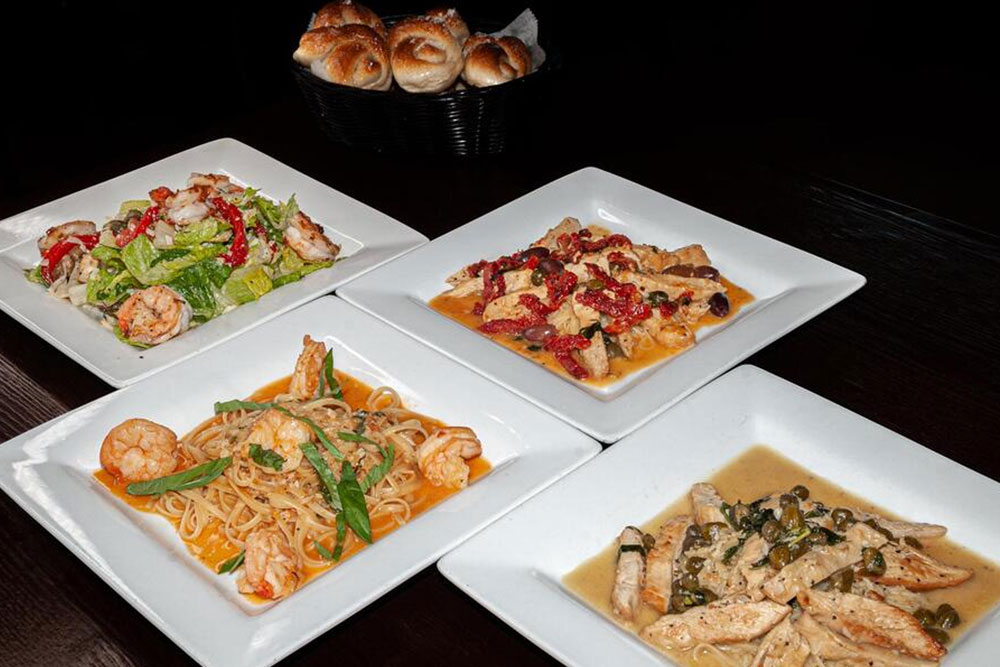 Four square white plates feature Italian dishes at Bella Sera Marble Falls: shrimp Caesar salad, creamy chicken pasta, shrimp pasta, and a chicken dish with capers, all on a dark table with a basket of rolls in the background.