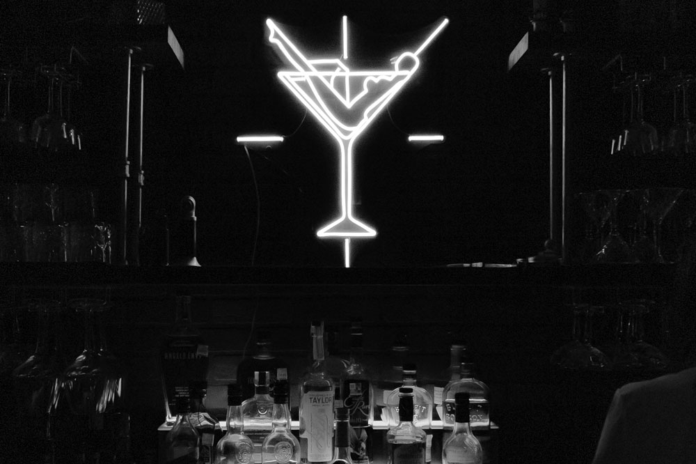 A glowing neon sign in the shape of a cocktail glass with an olive hangs above a bar shelf stocked with various bottles of liquor at Sips on Second in Marble Falls, set against a dark background.