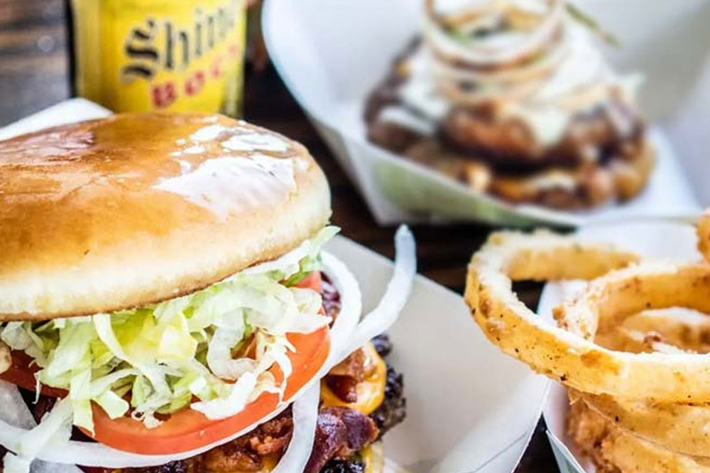 A close-up of a Bill's Burgers cheeseburger with lettuce, tomato, and onions, served with crispy onion rings. A bottle of Shiner Bock beer and another dish with fried food are in the background.