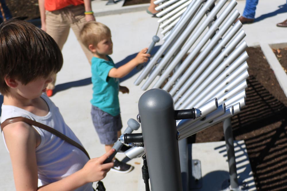 Two children play an outdoor xylophone with mallets on a sunny day at Harmony Park in Marble Falls. The xylophone’s silver metal bars form a graceful curve, while adults and other children enjoy the lively atmosphere in the background.