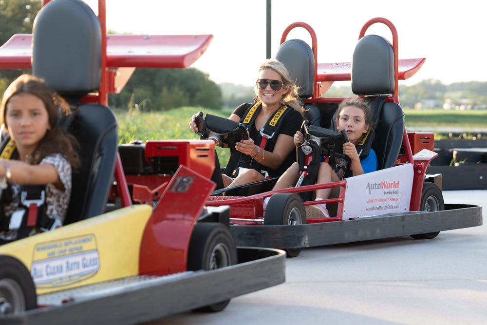 Three people enjoy go-karts at Putters and Gutters in Marble Falls. A woman and girl smile in a red kart, seatbelts fastened, while another girl drives a yellow kart up front. It’s a sunny day perfect for family fun.