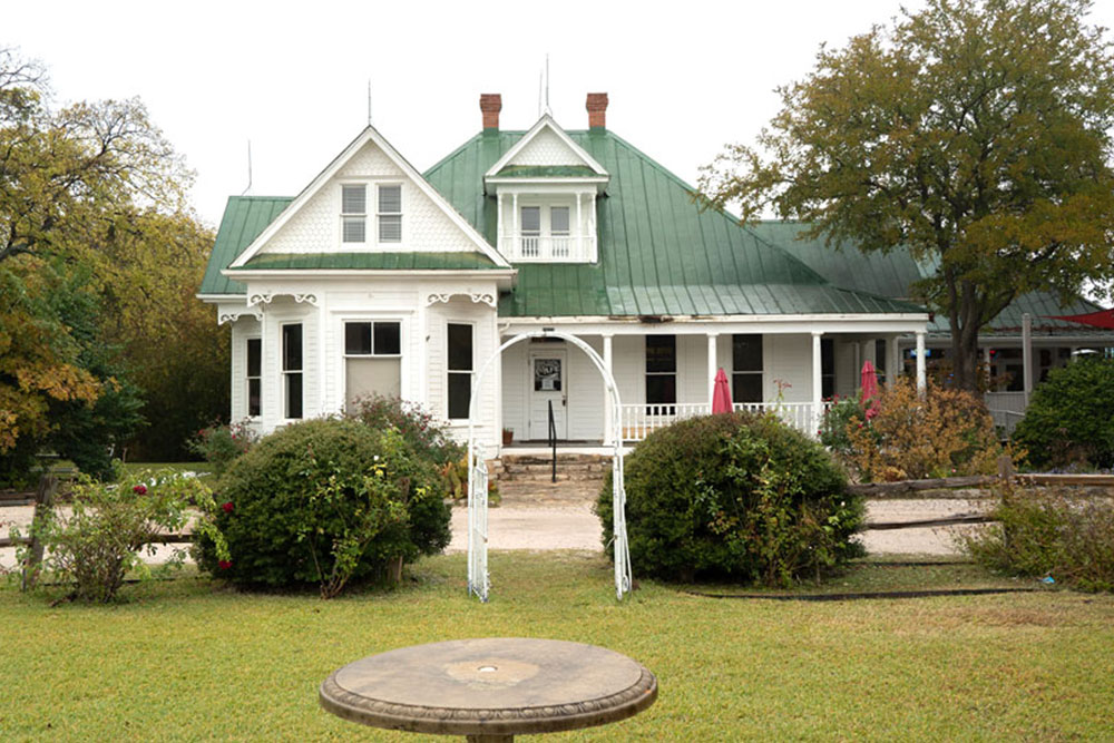 A large white Victorian-style house with a green metal roof, front porch, and steps leading up. This is Hooper's Kingsland house used in the filming of the original Texas Chainsaw Massacre movie, surrounded by bushes, trees, and a grassy lawn with a birdbath.