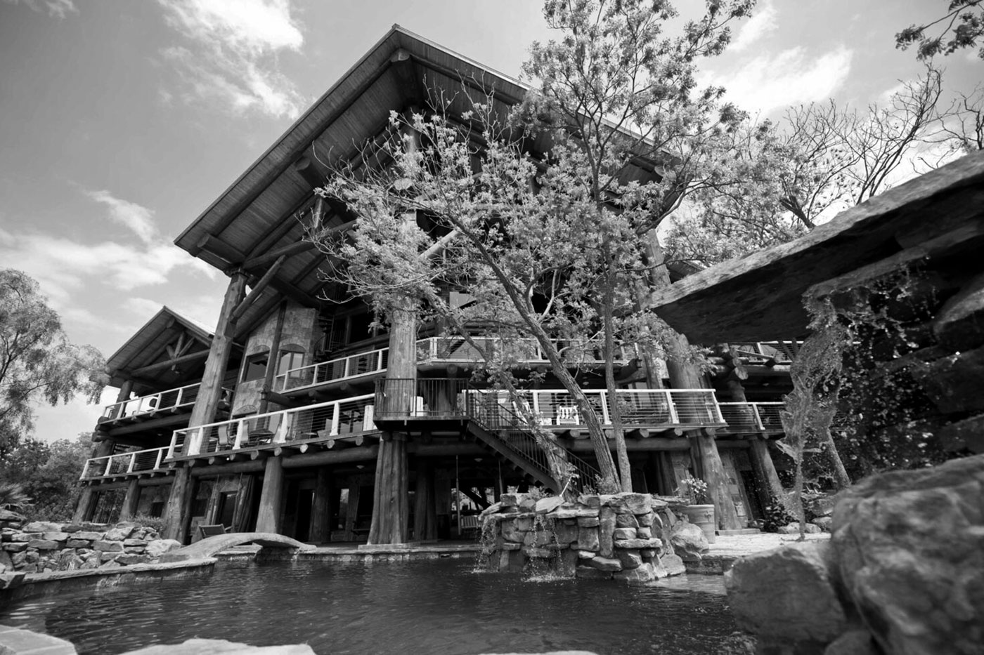 A large rustic log cabin with multiple stories, balconies, and exposed wooden beams stands by a pond with rocks and a small waterfall—an ideal Family Weekend retreat in scenic Burnet County, surrounded by trees under a partly cloudy sky.