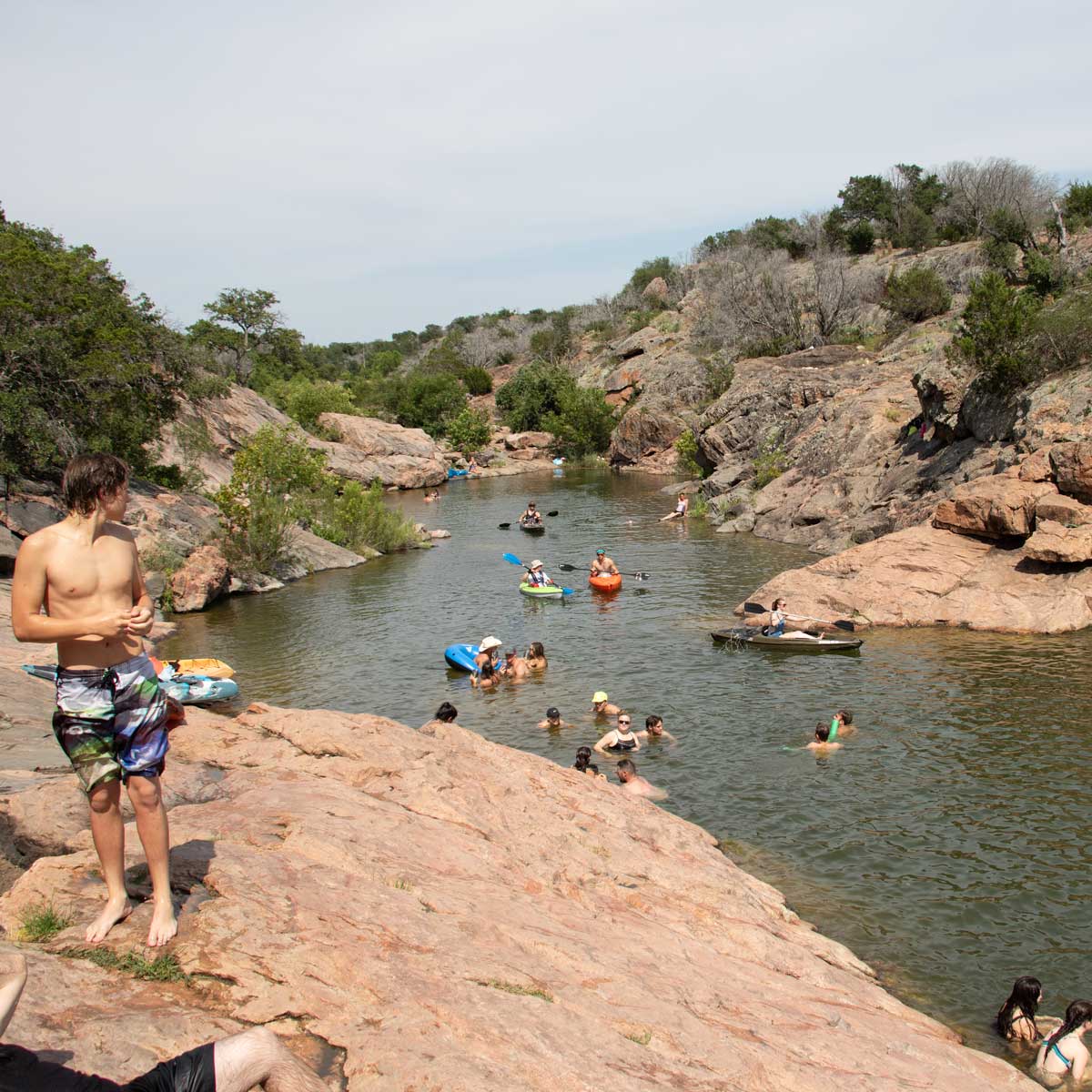 People swim, float, and relax in a rocky river surrounded by greenery. One person stands on the rocks in swim trunks, while others enjoy the water on floats or swimming. The sky is clear, suggesting a warm, sunny day.
