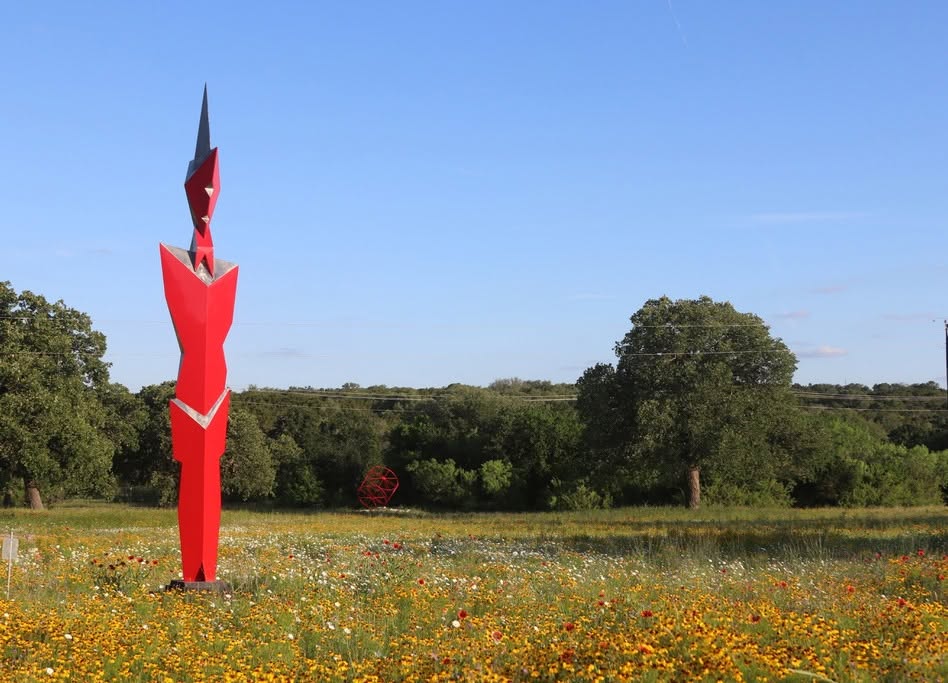 A tall, angular red sculpture—an impressive work of art—stands in a wildflower field at Museo Benini, with trees and another red artwork in the background under a clear blue sky.