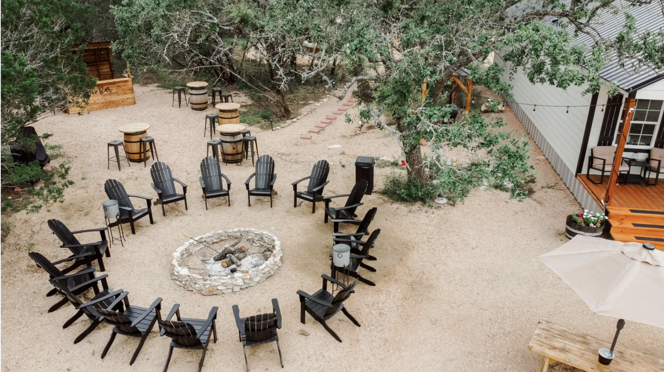 A circular arrangement of black chairs surrounds a stone fire pit in a gravel yard with trees, wooden tables, and a building with a deck nearby—creating an inviting setting perfect for gatherings at the Vital Reset Retreat.
