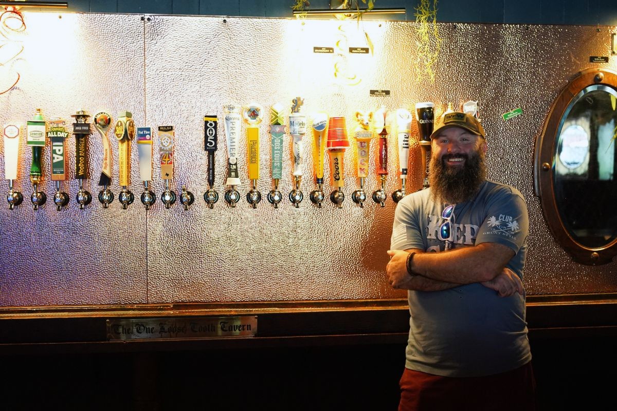 A man with a beard stands smiling in front of a wall lined with various beer taps in a dimly lit bar, ready to say cheers and grab a drink. A round mirror hangs on the wall to his right, reflecting the welcoming atmosphere of Highland Lakes.