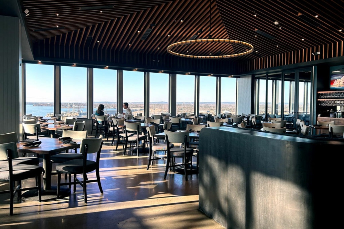 A modern restaurant with large windows, a wooden ceiling, circular chandelier, and mostly empty tables offers the perfect spot to grab a drink. Two people sit by the window enjoying drinks as sunlight streams across the polished floor—an inviting setting inspired by the relaxed charm of Highland Lakes.