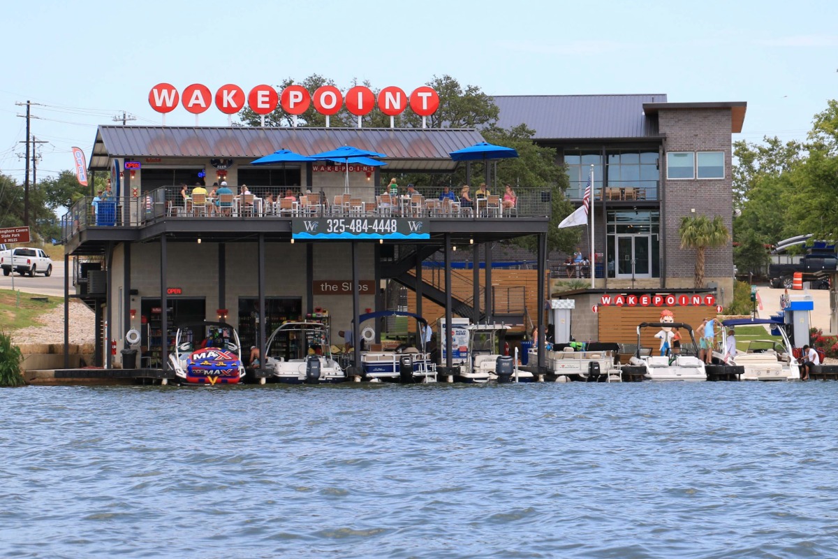 A two-story waterfront restaurant with boats docked below, people dining on an upper patio, and a red Wakepoint sign on the roof. The lively scene overlooks the beautiful Highland Lakes, where guests gather to grab a drink and cheers to good times by the water.