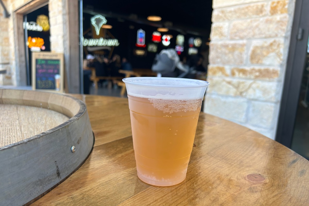 A plastic cup filled with light beer sits on a wooden table outdoors, near the entrance of a bar or restaurant with people and neon signs visible inside the building—perfect for those moments when you just want to grab a drink and say, “Cheers to here,” in Highland Lakes.
