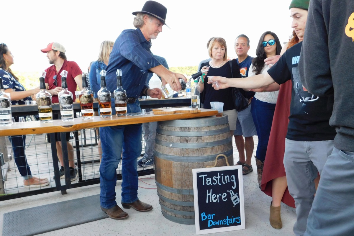 A man in a hat pours whiskey samples at an outdoor tasting event in the heart of Highland Lakes. Several people stand in line by a barrel with a sign that reads Tasting Here, Bar Downstairs, ready to grab a drink and enjoy the experience. Four bottles are displayed on the table—cheers to good company and great spirits!.