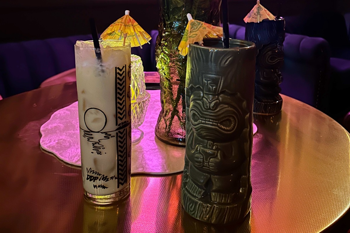 Three tropical drinks in tall tiki mugs with straws and paper umbrellas sit on a shiny round table, inviting you to grab a drink and say cheers, with colorful lighting and a decorative centerpiece in the background—perfect for relaxing by the Highland Lakes.