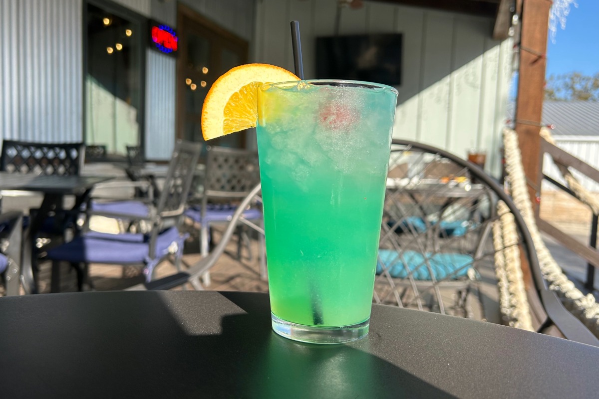 A bright blue-green cocktail with ice, an orange slice, and a cherry garnish sits on an outdoor table at Highland Lakes; patio chairs and a building are visible in the background—perfect for grabbing a drink and toasting “cheers” with friends.