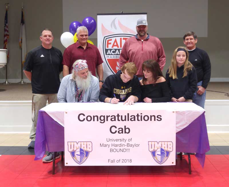 Faith Academy of Marble Falls senior football player Cab Booth (seated, middle) signs his national letter of intent to play for the University of Mary Hardin-Baylor as his parents, Jim and Angela Booth (seated), Faith Academy athletics director Randy Denton (standing, left), head football coach Pete Rhoades, former Flames head coaches Stephen Shipley and Russ Roberts, and sister, Seri, look on. Staff photo by Jennifer Fierro