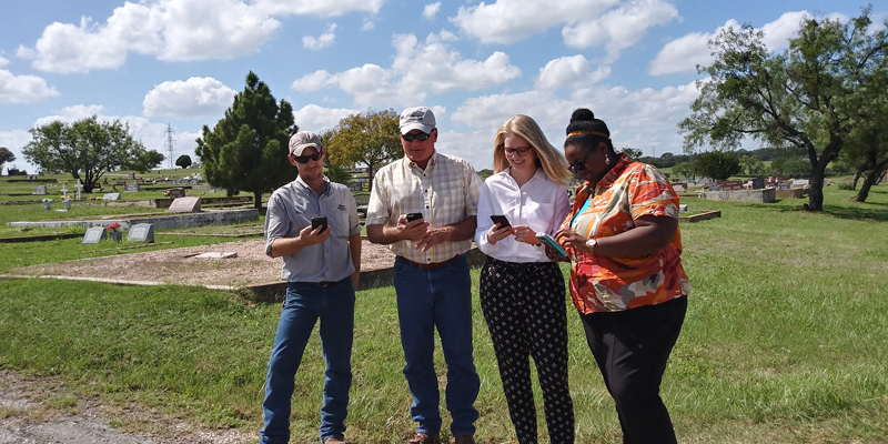 Brian Murphy (left) and Lewis Fletcher, both of the Marble Falls Parks and Recreation Department, Chelsea Seiter-Weatherford, GIS analyst for the city of Marble Falls, and Monique Breaux, administrative assistant of the Parks and Recreation and Water departments, are using their smartphones to pinpoint their exact location in Marble Falls City Cemetery through the city’s new online mapping system. Staff photo by Jennifer Fierro