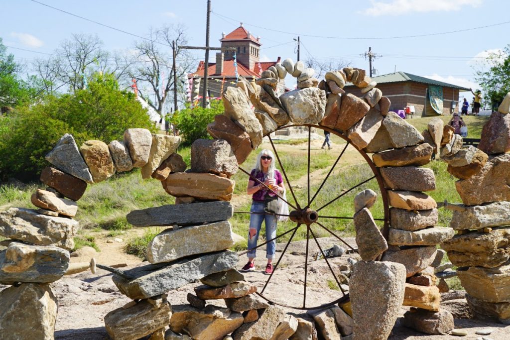 A woman stands behind a circular rock structure with metal spokes in an outdoor area, with a brick building and greenery in the background.