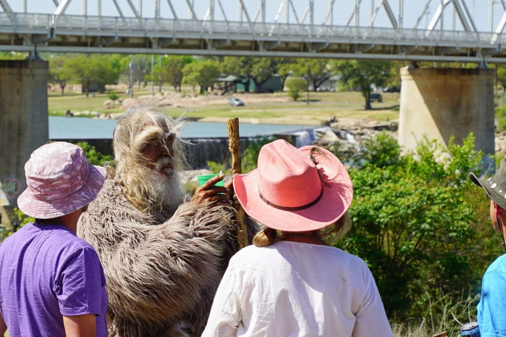 A person in a furry costume holding a staff talks to three people in hats near a river and bridge on a sunny day.